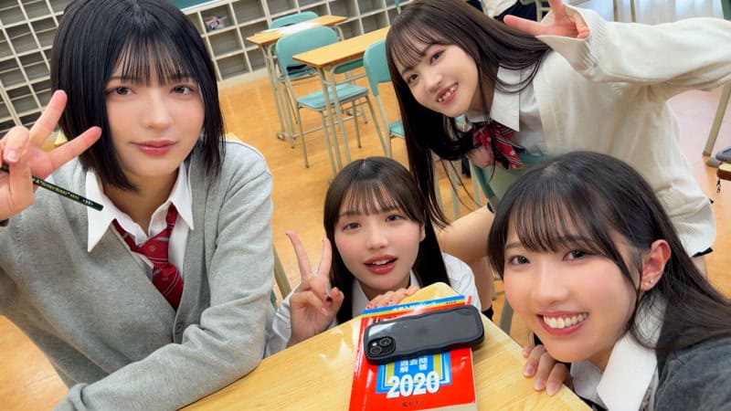 教室の机を囲む制服姿の4人が、参考書とスマホの前でカメラを見る